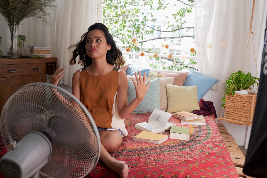 Asian girl in front of fan, thinking about calling air conditioning services to beat the heat. 