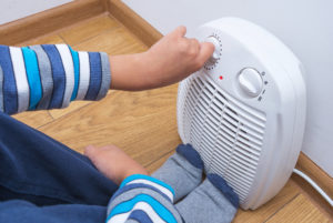A young boy warms himself near an electric fan heater, sitting on the floor at home. Part of body, selective focus.