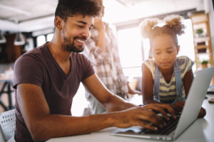 father working from home on his laptop as his daughter watches with the sun shining through the window